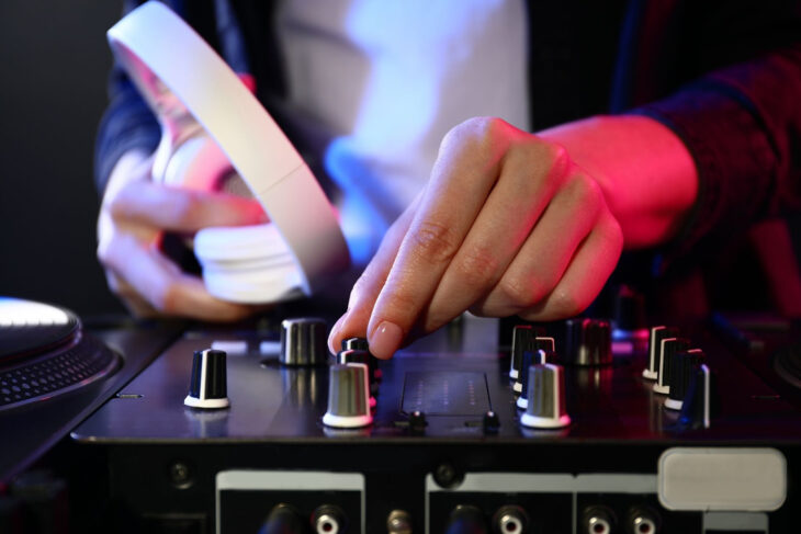 A female EDM artist holding white headphones while adjusting knobs on a mixer